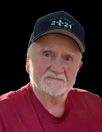 An older man with a white beard and mustache wears a navy "Mill Creek 2021 Fire" cap and a red shirt, looking into the camera against a solid black background.