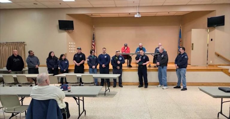 A group of people in uniform stand in a line at the front of a room, facing an audience seated at tables. Three people sit behind a desk on a raised platform in the background. An American flag is visible.