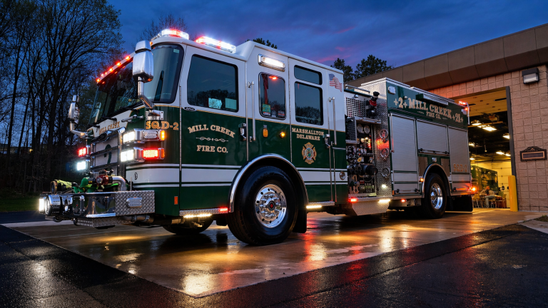 A green and white fire truck with flashing lights is parked outside a fire station at dusk, reflecting on the wet pavement. The truck is labeled "Mill Creek Fire Co. 2-4 Mill Creek" and "Marshallton Delaware.