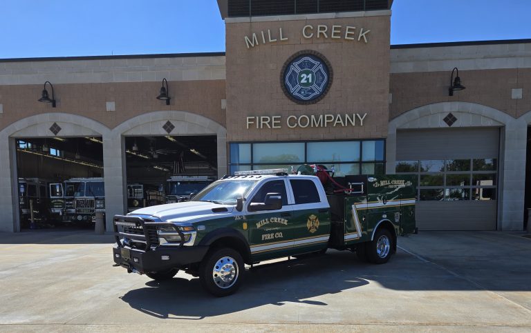 A green and white Mill Creek Fire Company fire truck is parked in front of the Mill Creek Fire Company station, with the station’s sign, trucks, and garage bays visible in the background.