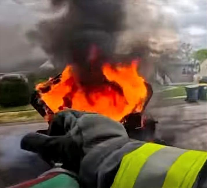A firefighter in protective gear aims a hose at a car with a hood engulfed in bright orange flames on a suburban street.