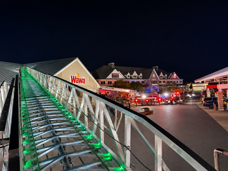 At night, emergency vehicles with flashing lights are parked outside a Wawa store and other buildings, seen from atop a firetruck ladder illuminated by green lights. People are gathered near the scene.