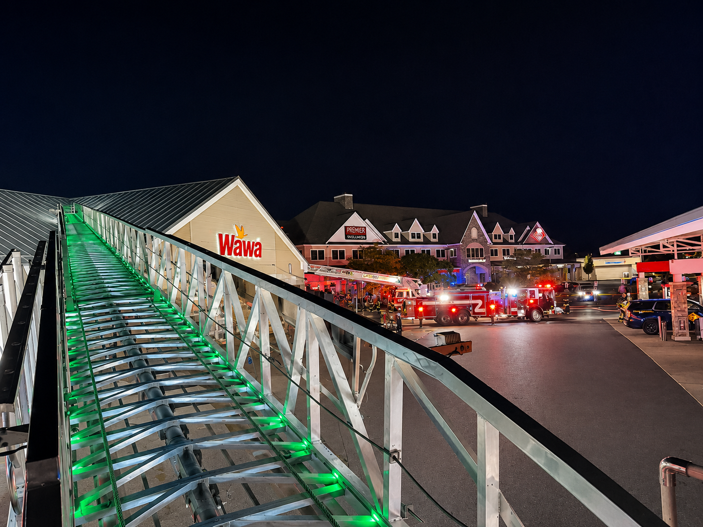 At night, emergency vehicles with flashing lights are parked outside a Wawa store and other buildings, seen from atop a firetruck ladder illuminated by green lights. People are gathered near the scene.