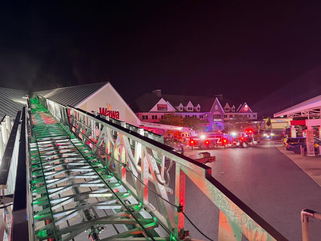 A nighttime scene shows fire trucks and emergency vehicles with flashing lights outside a Wawa store. A large fire truck ladder extends toward the building. Several responders are present.