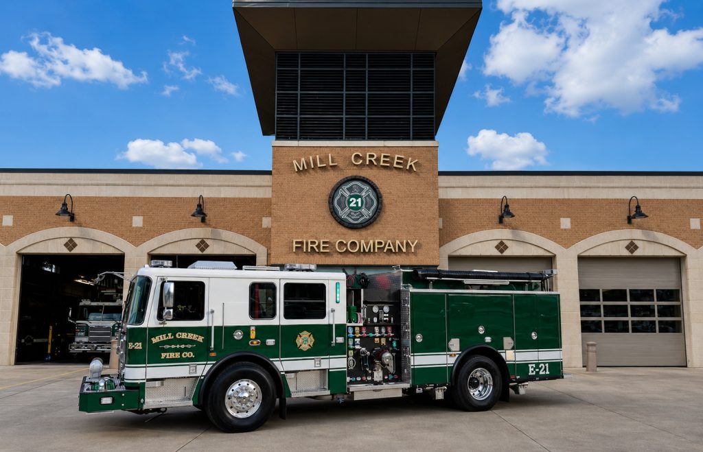A green and white fire truck is parked in front of the Mill Creek Fire Company building, which has three garage doors and a large badge with “21” above the main entrance. The sky is partly cloudy.