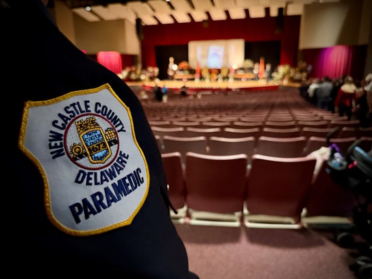 Close-up of a New Castle County, Delaware paramedic uniform patch in the foreground, with an auditorium filled with rows of empty chairs and a stage with people and a projection screen in the background.