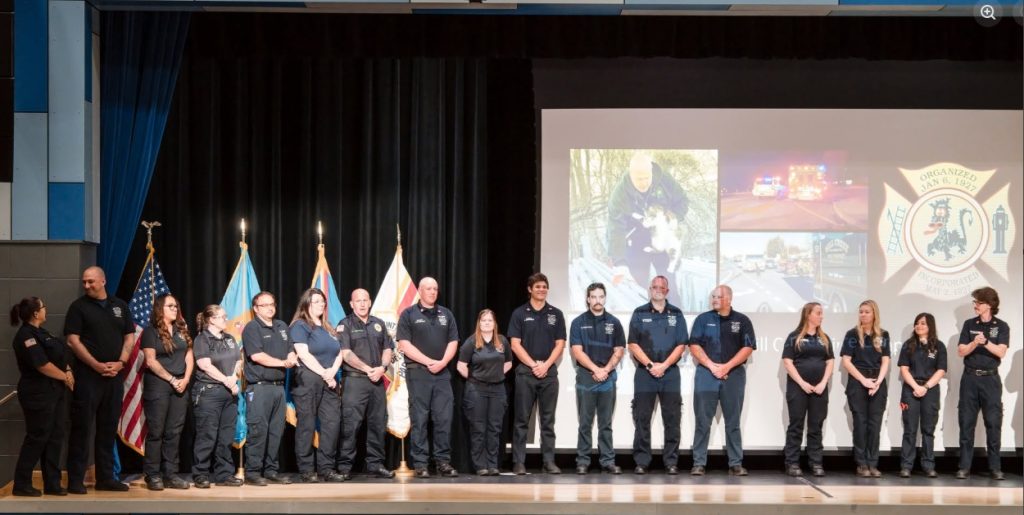 A group of uniformed emergency responders stands on stage in a line, with flags behind them and a large screen displaying various images and a badge emblem in the background.