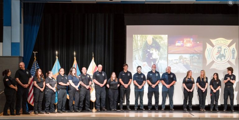 A group of uniformed emergency responders stands on stage in a line, with flags behind them and a large screen displaying various images and a badge emblem in the background.