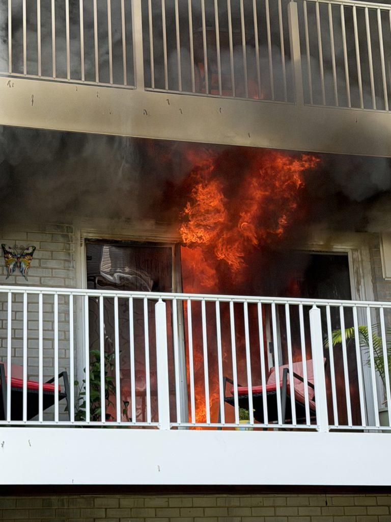 Bright flames and thick black smoke pour out of a window behind a white balcony railing, with chairs and plants visible on the balcony.