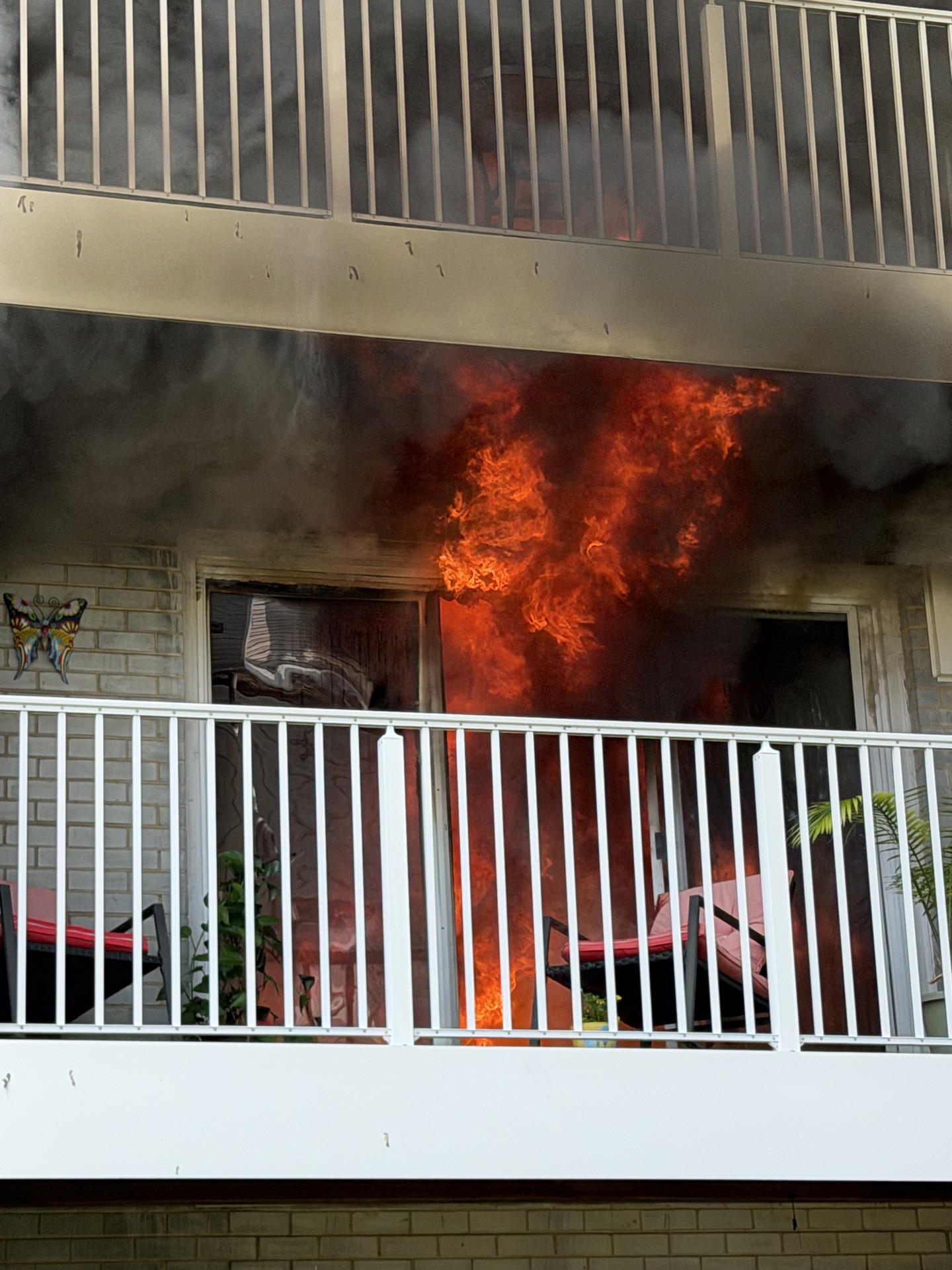 Bright flames and thick black smoke pour out of a window behind a white balcony railing, with chairs and plants visible on the balcony.