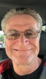A middle-aged man with gray hair, glasses, and a slight smile sits in a car, wearing a black and red shirt.