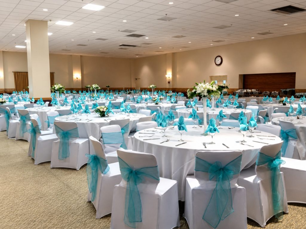 Large banquet hall with round tables covered in white linens, each decorated with turquoise chair sashes and napkins. Elegant flower centerpieces are on the tables, all set for a formal event.