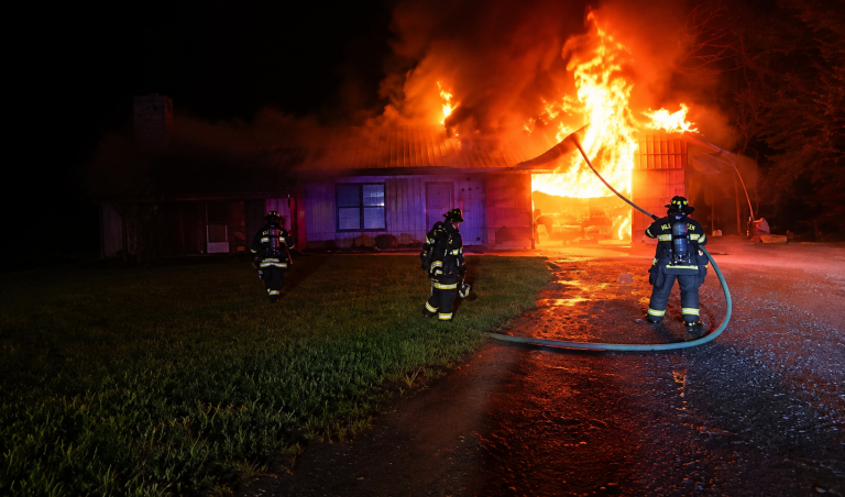 Three firefighters work to extinguish a large blaze engulfing the roof of a house at night, with flames and smoke billowing into the dark sky.