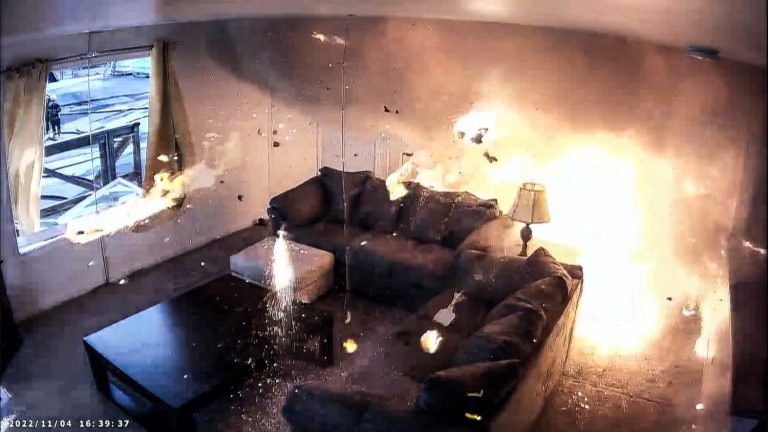 A living room with a sofa and table is engulfed in flames and smoke, debris flying through the air, suggesting an explosion has just occurred.